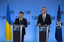 Two men in business suits stand at silver podiums, with a blue and yellow Ukrainian flag to their left and a blue and white NATO flags their right.