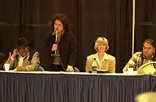 Lanscape photograph displaying woman with microphone speaking and 3 other individuals sitting next to her. Susan Masten, standing, with Secretary Gale Norton (right of Masten) and Three Affiliated Tribes Chairman Tex Hall (right of Norton)