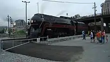 People standing next to a black streamlined steam locomotive in the foreground. The railroad signals, road bridge, and buildings are located next to the locomotive in the background