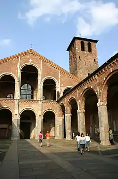 The facade and forecourt of a redbrick church are composed of simple arcades. A brick tower rises up to one side.