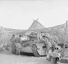 Three men work on a tank, which is under a net and facing to the left.