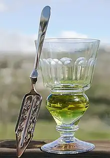 stemmed reservoir glass containing a green-coloured liquid and a flat, slit, absinthe spoon