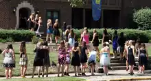 Several dozens of college women in casual dresses line up on the stairs leading through a lawn in front of a sorority house (Alpha Xi Delta).