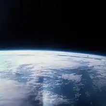 The edge of Earth, heavily overexposed, in the lower half of the image, with black space above. In between them, a blue haze layer from the atmosphere. There is a lens flare in one corner.
