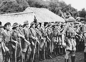 Soldiers on parade in front of a hut in a tropical setting. An officer in a steel helmet with a walking stick stands in front facing away from them, while the men behind him are wearing a various assortment of uniforms including steel helmets, slouch hats, shorts and are carrying rifles