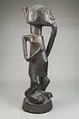 Figurine of a standing woman; late 19th or early 20th century; wood; 27.9 × 8.3 × 10.2&nbsp;cm (11 × 3.mw-parser-output .frac{white-space:nowrap}.mw-parser-output .frac .num,.mw-parser-output .frac .den{font-size:80%;line-height:0;vertical-align:super}.mw-parser-output .frac .den{vertical-align:sub}.mw-parser-output .sr-only{border:0;clip:rect(0,0,0,0);clip-path:polygon(0px 0px,0px 0px,0px 0px);height:1px;margin:-1px;overflow:hidden;padding:0;position:absolute;width:1px}1⁄4 × 4 in.); Brooklyn Museum (New York City)