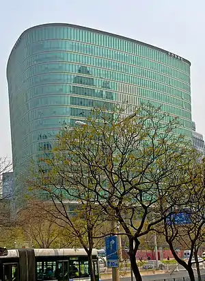 A triangular greenish glass-faced building, flaring outward the higher it gets, rises over a street lined with freshly budding trees on which a blue and white trolleybus is stopped