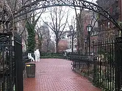 A color photograph of Christopher Park in winter, showing the wrought iron entrance arch in the foreground and the brick pavement surrounded by five and six story brick buildings; in the center background are four white statue figures: two males standing, one with his hand on the other's shoulder and two females seated on a park bench, one woman with her hand touching the other's thigh. All are dressed in jeans and loose clothing