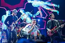 Two men play their guitars as one in the middle sings with a microphone. Meanwhile, a fourth man is playing drums behind the other three.