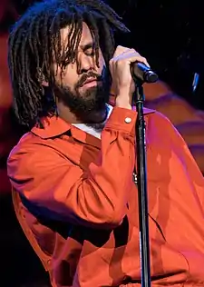 J Cole singing with his eyes closed while wearing an orange jacket and holding onto his microphone stand.