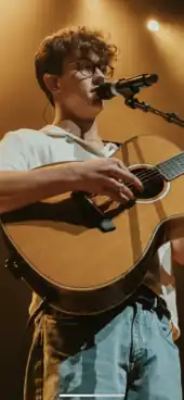 A man with curly hair playing a guitar and singing into a microphone while standing