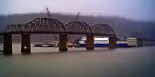 The bridge has 6 spans; the 5th has been removed from the bridge.  Underneath, the ship Delta Mariner is three quarters of the way through the bridge, with the destroyed 5th bridge span draped over its front.  Two boats with cranes sit at the bow of the Delta Mariner.