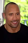 A man with dark skin, and closely shaven hair, looks to his left slightly, while smiling, and wearing a black shirt.