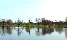 Meadow Lake with baseball fields behind it.  Interstate 95 borders the park, with the Philadelphia Naval Shipyard behind it. Above an airplane begins its landing at Philadelphia International Airport.