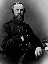 A bearded man with his right hand in his jacket and his left on a table.