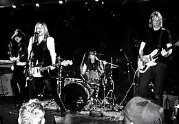 A five-piece rock band performing live on stage. A red sign behinds them reads "SPIN 20" in white lettering.