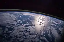 Image 16A view of Earth with different layers of its atmosphere visible: the troposphere with its shadows casting clouds and a band of stratospheric blue sky at the horizon, and above this a line of green airglow of the lower thermosphere around an altitude of 100 km, at the edge of space. (from Earth)