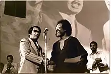 Jim Jones shakes hands with Cecil Williams with a large picture of Martin Luther King Jr. in the background.