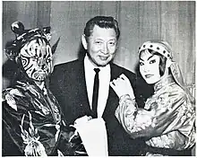 Black and white photograph of Johnny Kan, a restauranteur in Chinatown, San Francisco. Kan is wearing a dark suit with a dark tie. He stands between two actors, a devil on the left side and an angel on the right side, who belong to the Foo Hsing Opera Company.