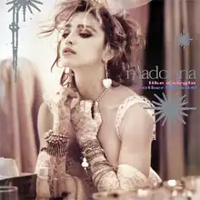 Madonna wearing a wedding dress and heart-shaped earrings is using one of her gloved hands to pull down on her pearl necklace.