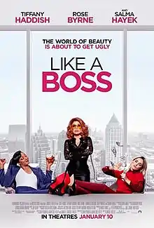 A red-haired woman wearing black outfit stands with her arms crossed at a boardroom table, behind her is view of the city. In front sit two woman with their feet up on the table, one dressed in blue, the other in red.
