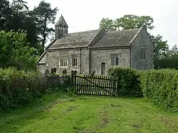 A small stone church seen from an angle; behind the chancel is a slightly higher nave with a bellcote at the far end. A porch protrudes beyond that