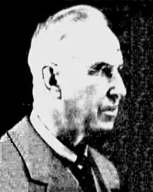 A black-and-white photographic portrait of an older Costello from the shoulders up, wearing a suit and eyeglasses