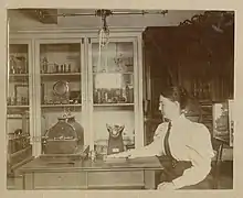 While sitting, Mabel Augusta Chase places her hand on a glass photographic plate which sits on a table below a crookes tube to take a radiograph of the bones in her hand. She is in the Wellesley physics laboratory.