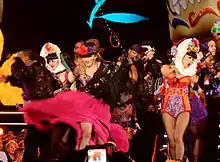 Picture of a group of people dressed in colorful, Mexican-styled clothing. The woman in the middle is blonde, and is wearing a black shawl, black hat, and pink skirt.