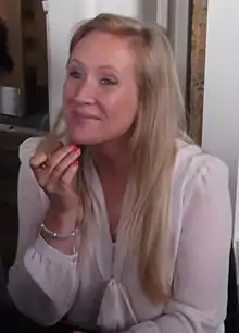 White woman with blonde hair smiling at someone with her left hand resting on her chin.
