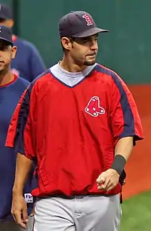 A white male baseball player with facial hair, wearing a blue cap with a red "B" on it, a red polyester shirt, and white pants, looks to his left.