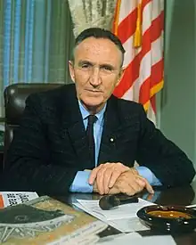 A color photo of Mansfield sitting at his desk in his office, smiling, in 1966.