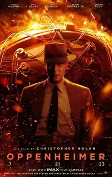 J. Robert Oppenheimer in a hat and suit stands in front of the nuclear bomb.