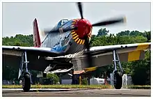 A single nosed-propeller aircraft is on the ground on its wheels with the propeller in motion. The aircraft is viewed from the front, but the red nose faces slightly to the right. The aircraft has black propellers. Parts of the wings and propellers are cropped from view.