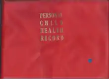 The front cover of a book, with a solid red background and the words 'PERSONAL CHILD HEALTH RECORD' in gold serif justified typeface on alternating lines. A small blue circular sticker is present in the bottom right.