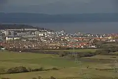 Multiple buildings including terraces, detached houses and blocks of flats. In the foreground are fields and in the background water and then hills.