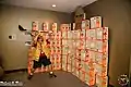 A wall of empty Maruchan ramen noodle boxes with a cardboard figure from Attack on Titan being swatted at by a member of Anime Midwest's staff.