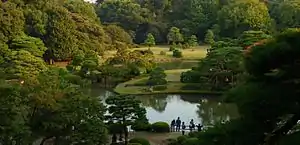 A panoramic view of the Rikugi-en from the Fujishiro-toge hill vantage point. Green trees surround a serene lake.