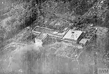Black and white aerial photo of a multi-story building with a tall mast next to it. Smoke is visible in the air in front of the building.