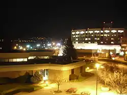 The photo was taken during the night. A large building in the foreground is brightly lit, with a much larger building behind it, and city lights in the background