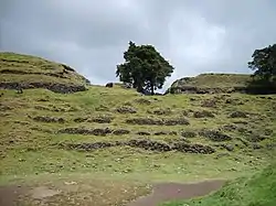 A series of semi-collapsed dry-stone terraces, overgrown with short grass. On top of the uppermost of five terraces stand the crumbling, overgrown remains of two large buildings flanking the ruins of a smaller structure. A tree grows from the right hand side of the smaller central building, and another stands in at extreme right, on the upper terrace and in front of the building also standing on it. The foreground is a flat plaza area, with the collapsed flank of a grass-covered pyramid at bottom right. The sky is overcast with low rainclouds.