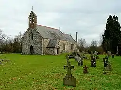 A stone church seen from the southwest, with a slate roof and red tiled ridge. At the west end is a door a window and a double bellcote; protruding from the south side is a porch