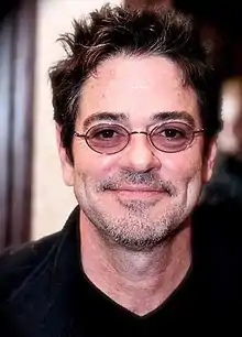 A man with short brown hair and purple-tinted glasses smiles towards the camera.