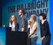 Four smiling people (three women and one man) stand in front of a podium. Behind them is a large blue screen with the words THE FULLBRIGHT COMPANY projected across it.