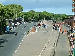 alt=TT
Grandstand area with scoreboard to extreme left, course start/finish to left (in distance), pit lane with re-fuelling equipment and acceleration lane to centre