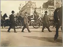 Several men walk down the road. Roosevelt sits in a horse drawn carriage. A large crowd watches in the distance.