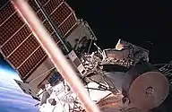 A man dressed in a spacesuit seen crawling along a white, cylindrical space station module. A large solar array can be seen projecting from the top of the module, and various other pieces of apparatus are visible. The Earth's horizon and space are visible behind the solar array.