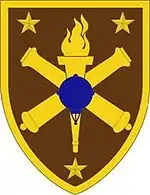 A brown shield shaped patch with a yellow border. Yellow stars are in each corner, surrounding crossed yellow cannons superimposed by a torch in yellow, which in turn is superimposed by a blue sea mine