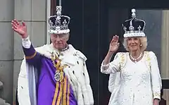 Charles and Camilla wearing their crowns and coronation robes waving from the balcony of Buckingham Palace