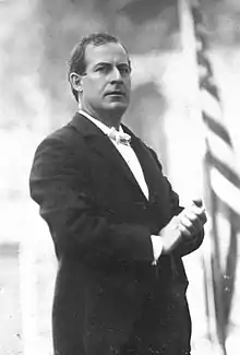 A man in his thirties wearing a dark suit holds his hands together in front of him as he looks to his right. In the background can be seen the stars and stripes.